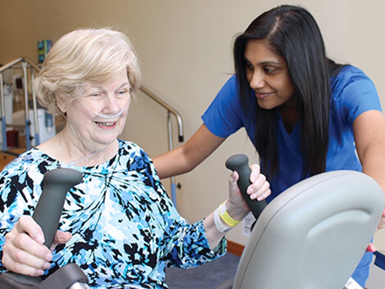 An elderly female patient using an elliptical machine, with assistance from a therapist.
